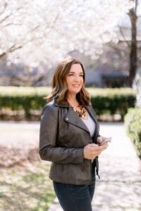 Smiling woman in a leather jacket holding a phone outdoors on a sunny day, with blooming trees and a residential neighborhood softly blurred in the background.