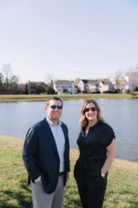 Two smiling real estate professionals standing by a lakeside neighborhood in North Atlanta on a sunny day, with homes in the background