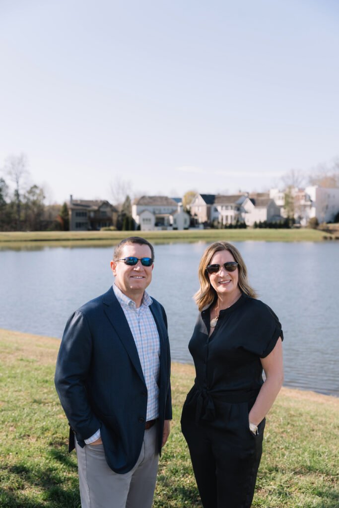 Two smiling real estate professionals standing by a lakeside neighborhood in North Atlanta on a sunny day, with homes in the background